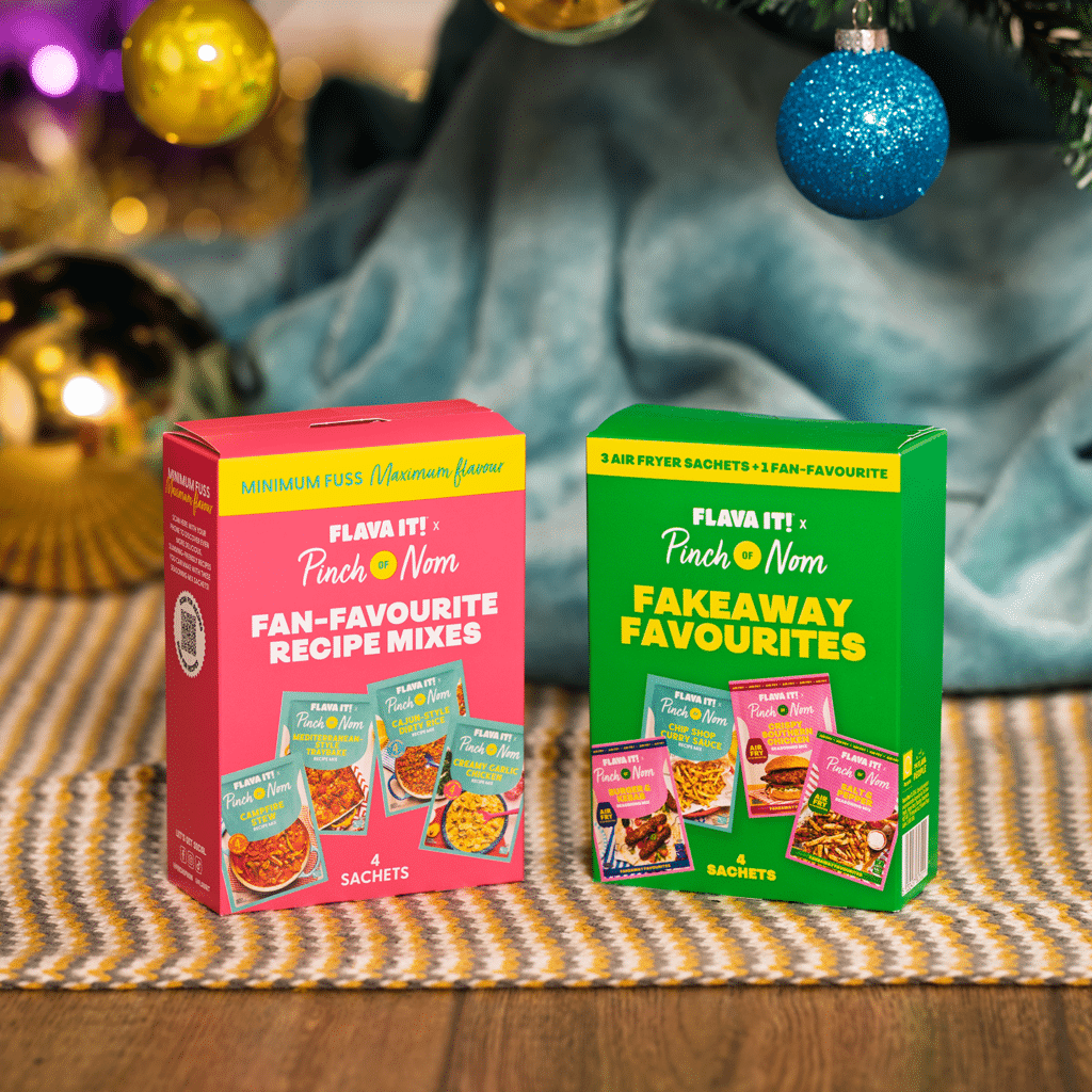 Two Pinch of Nom x Flava It! sachet collection boxes stand under a colourfully decorated Christmas tree, as though it has just been unwrapped on Christmas morning.