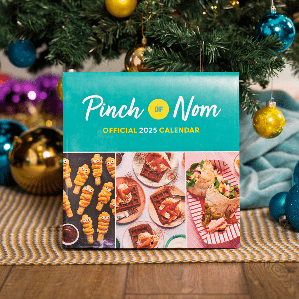 Pinch of Nom's official 2025 calendar stands under a colourfully decorated Christmas tree, as though it has just been unwrapped on Christmas morning.