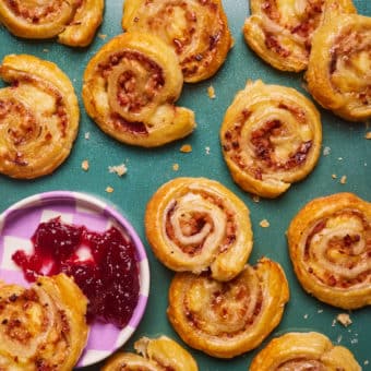 A freshly-baked batch of Pinch of Nom's slimming-friendly Bacon, Brie and Cranberry Swirls are scattered across a green countertop; a half-eaten swirl is being dipped into a small pot of cranberry sauce.