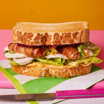 Pinch of Nom's slimming-friendly Boxing Day sandwich is served on a colourful countertop against a bright yellow backdrop. The filling of pigs in blankets, lettuce, turkey, sriracha-mayo, cranberry sauce and homemade festive slaw is visible at the centre of the sandwich.