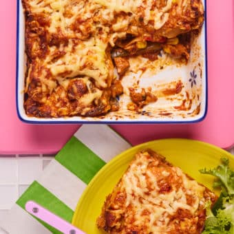 On a bright pink tabletop, Pinch of Nom's slimming-friendly Chicken Fajita Lasagne is served in a white oven dish. A portion has been scooped out onto a yellow plate, with a leafy green side salad and a fork waiting to tuck in.