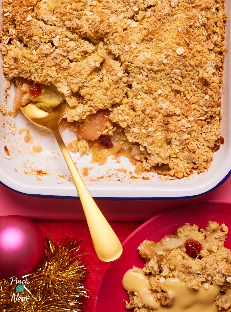 Pinch of Nom's Christmas Crumble is served in a white oven dish, set on a pink place mat. The table is set for Christmas, and a gold serving spoon rests in the dish. A portion has been served onto a small red plate nearby.