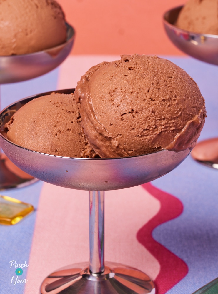 A table is placed against a light pink wall, topped with a funky-striped tablecloth. On the table, 3 portions of Pinch of Nom's low-calorie Ninja Creami Hidden Veg Ice Cream are served in silver dessert dishes. Spoons wait to tuck in.