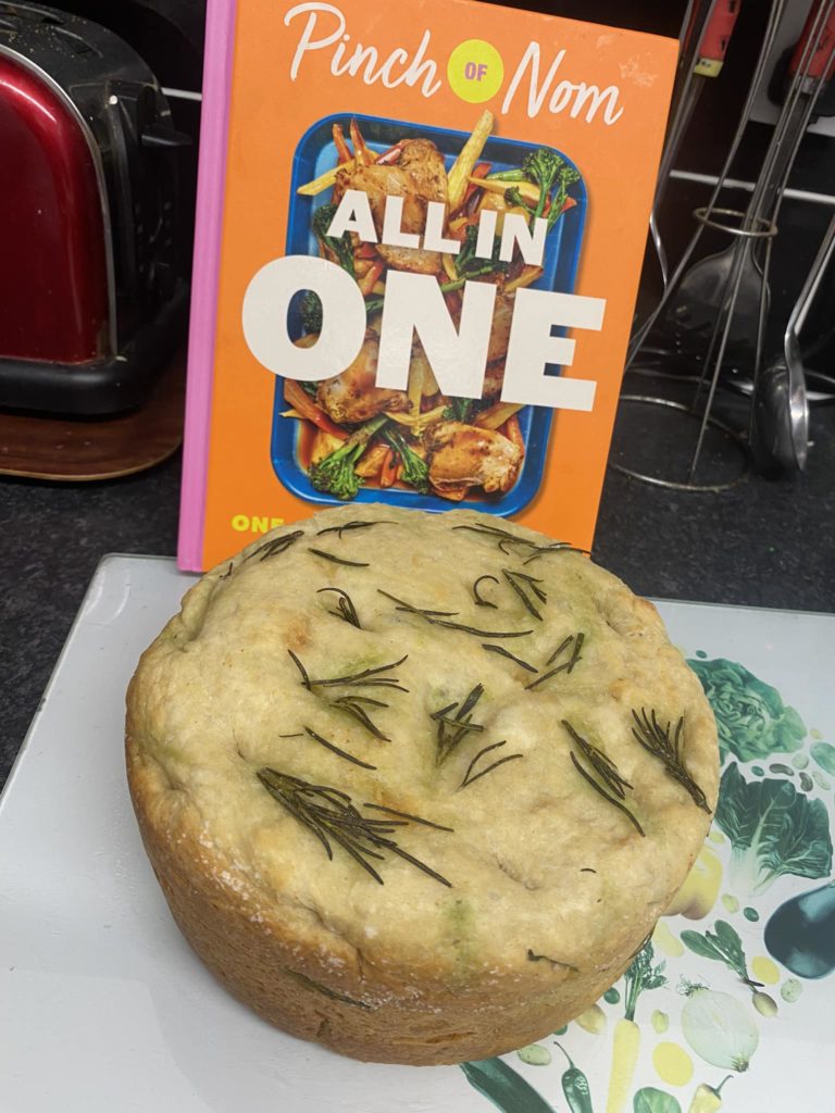 Vicky Galvin's Slow Cooker Focaccia is served on a white serving board, in front of a copy of Pinch of Nom: All in One.