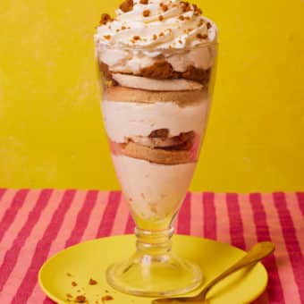 On a bright-pink striped tabletop against a yellow background, Pinch of Nom's slimming-friendly Biscoff Trifle recipe is served in a tall serving dish, topped with cream and crumbled biscuits. A spoon waits to dive in.