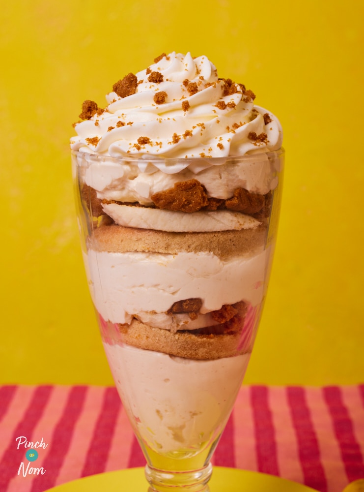 On a bright-pink striped tabletop against a yellow background, Pinch of Nom's slimming-friendly Biscoff Trifle recipe is served in a tall serving dish, topped with cream and crumbled biscuits.