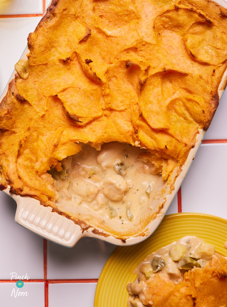 Pinch of Nom's slimming-friendly Chilli Chicken Pie with Sweet Potato Topping is served in a large ovenproof dish at the centre of a tiled worktop. A slice of the pie has been dished out onto a yellow plate, revealing the creamy filling inside.