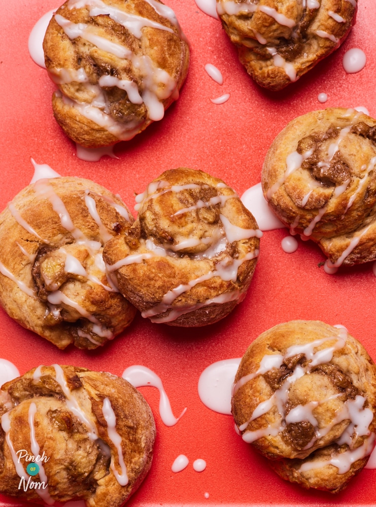 On a bright-red tray, a freshly-baked batch of Pinch of Nom's Banana Bread Cinnamon Rolls are laid out, drizzled with a generous amount of icing sugar. The sweet banana and cinnamon filling is bursting from the centre of each roll.