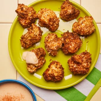 On a bright green plate, a freshly-baked golden brown batch of Pinch of Nom's slimming-friendly Air Fryer Cauliflower Cheese Florets are served. A small bowl is to one side, filled with the cheesy, homemade dipping sauce – ready to dig in!