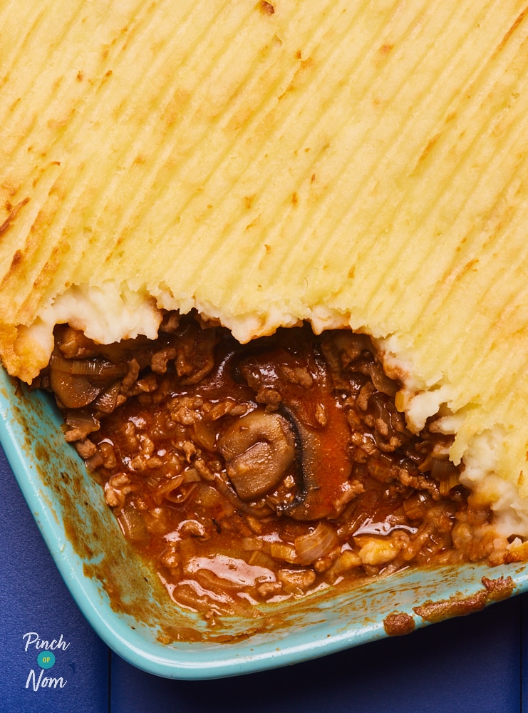 Pinch of Nom's slimming-friendly Garlic and Mushroom Cottage Pie is served in an oven dish; a portion has been removed and served, revealing the garlicky, mince filling inside.