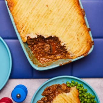 In a large ovenproof dish, Pinch of Nom's slimming-friendly Garlic and Mushroom Cottage Pie is served fresh from the oven at the centre of a table. A portion is plated up to to one side with a garden peas and salt and pepper mills.