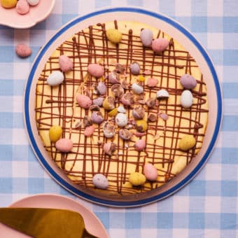 At the centre of a gingham table, Pinch of Nom's slimming-friendly Mini Egg Cheesecake is served decorated with a chocolate drizzle and a garnish of mini eggs, ready to be sliced.