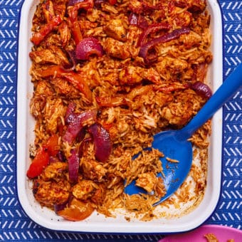 In an oven dish, Pinch of Nom's slimming-friendly Smoky Chicken and Rice Traybake is served at the centre of a table. A large blue serving spoon is dishing a portion out onto a pink side plate.