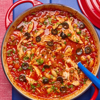 In a large red casserole pan, Pinch of Nom's slimming-friendly Spanish-Style Pulled Chicken is served at the centre of a table, with a large serving spoon waiting to begin dishing out. The freshly-made batch is packed with colour from lots of chopped vegetables, vibrant tomatoes and black olives.