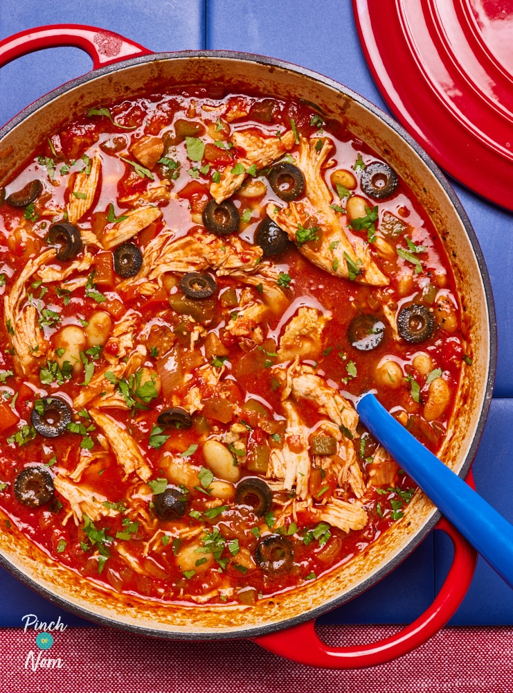 In a large red casserole pan, Pinch of Nom's slimming-friendly Spanish-Style Pulled Chicken is served at the centre of a table, with a large serving spoon waiting to begin dishing out. The freshly-made batch is packed with colour from lots of chopped vegetables, vibrant tomatoes and black olives.