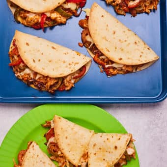 Four of Pinch of Nom's slimming-friendly Chicken Tacos are served filled with shredded chicken, peppers, and melted cheese on a blue baking tray, with three more tacos served on a bright green plate below.