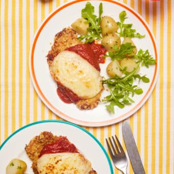 A table is set with two plates of Pinch of Nom's slimming-friendly Pork Parmigiana recipe. The cheesy-topped escalopes are served beside a green salad and new potatoes, with a set of cutlery to one side ready to dig in.