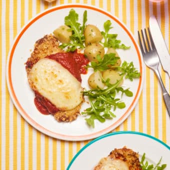 A table is set with two plates of Pinch of Nom's slimming-friendly Air Fryer Pork Parmigiana recipe. The cheesy-topped escalopes are served beside a green salad and new potatoes, with a set of cutlery to one side ready to dig in.