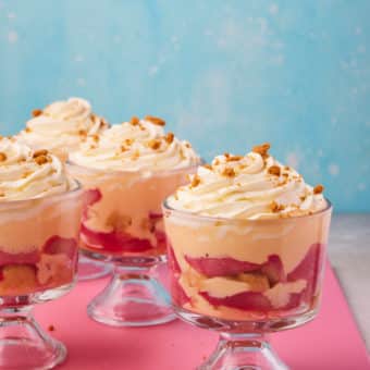 A table is set with four dessert dishes, filled with Pinch of Nom's slimming-friendly Rhubarb and Custard Trifles. The trifles are served in clear dessert glasses, revealing the bright-colours layers of yellow custard and pink rhubarb.
