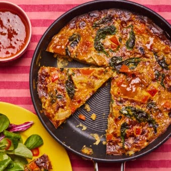 At the centre of a striped pink tabletop, a large frying pan is filled with Pinch of Nom's slimming-friendly Tikka-Style Frittata. A plate to one side holds a slice with a mixed salad.