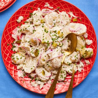 A table is set with Pinch of Nom's slimming-friendly Pickle Potato Salad served on a bright red plate, seasoned with salt and pepper, with finely sliced chives sprinkled on top.
